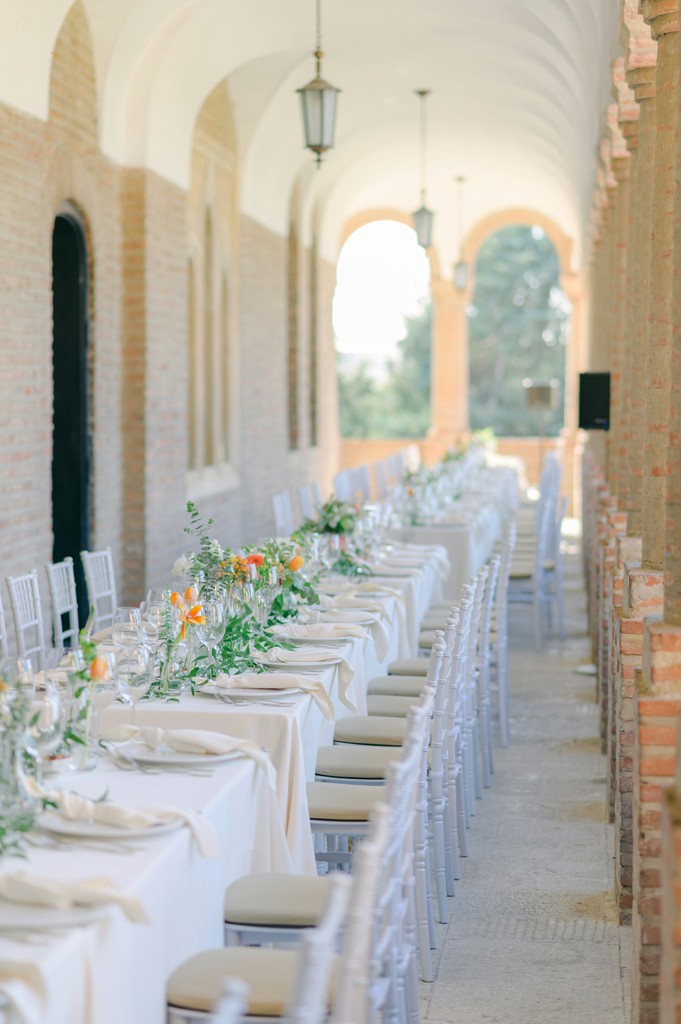 Elegant wedding reception table set with white linens and greenery under a sunlit brick archway at a Mediterranean-style venue
