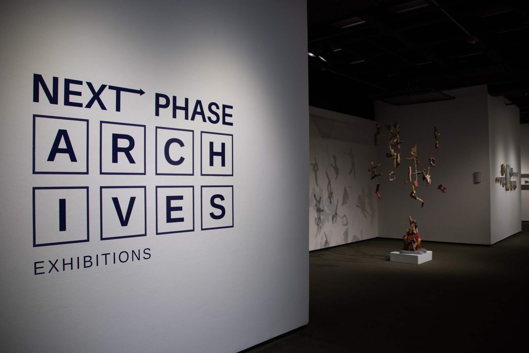 A label for ARCHIVES: Next Phase on a gallery wall with an installation of painted hanging wood pieces by Julie Glass around the corner