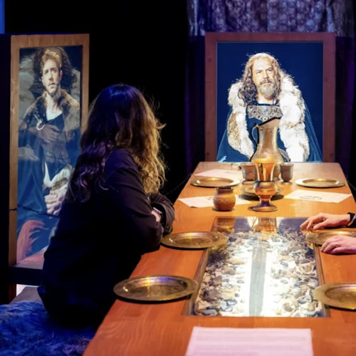 两个人坐在一个中世纪风格的桌子旁，桌上摆放着酒杯和盘子，面前是历史人物的真人大小肖像画。
