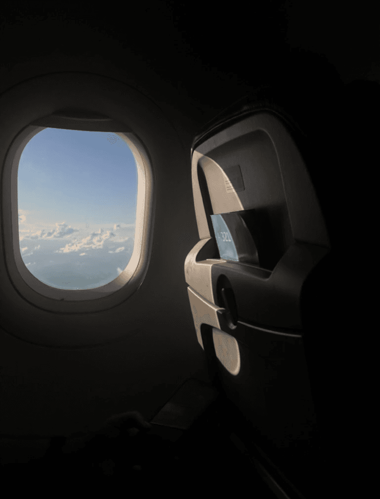Janela de avião vista de dentro da cabine, mostrando o céu com nuvens iluminado pela luz externa.
