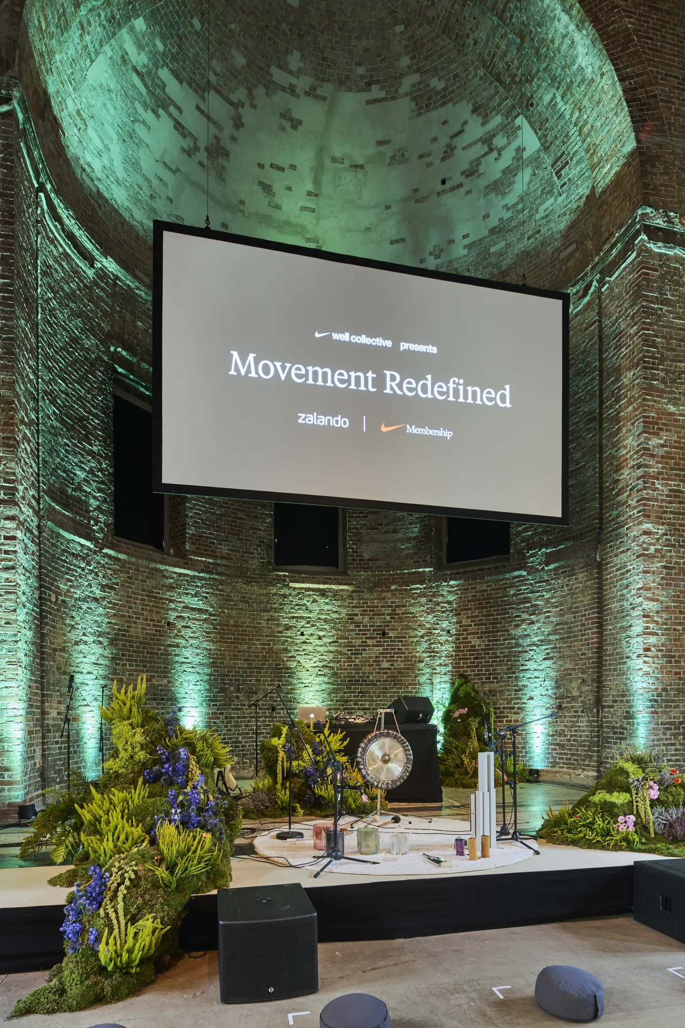 Stage in a historic brick hall showing a large LED screen with „Movement Redefined" branding by Zalando and Nike, framed by a lush moss-and-floral installation and seating cushions.