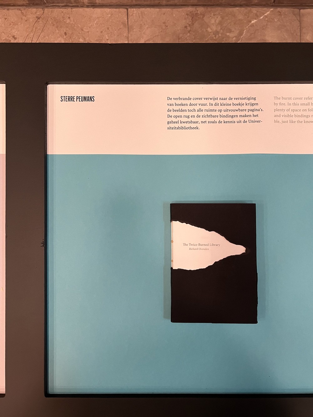 Een presentatie van het boek op de tentoonstelling. Het boek ligt op een tafel tegen een blauwe achtergrond, vergezeld van tekst en uitleg over de specifieke keuzes in het grafisch ontwerp.