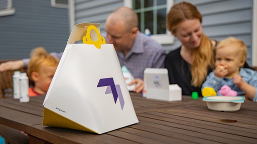 A family with two small children enjoys a meal outdoors on a wooden table, featuring branded takeaway packaging prominently in the foreground.