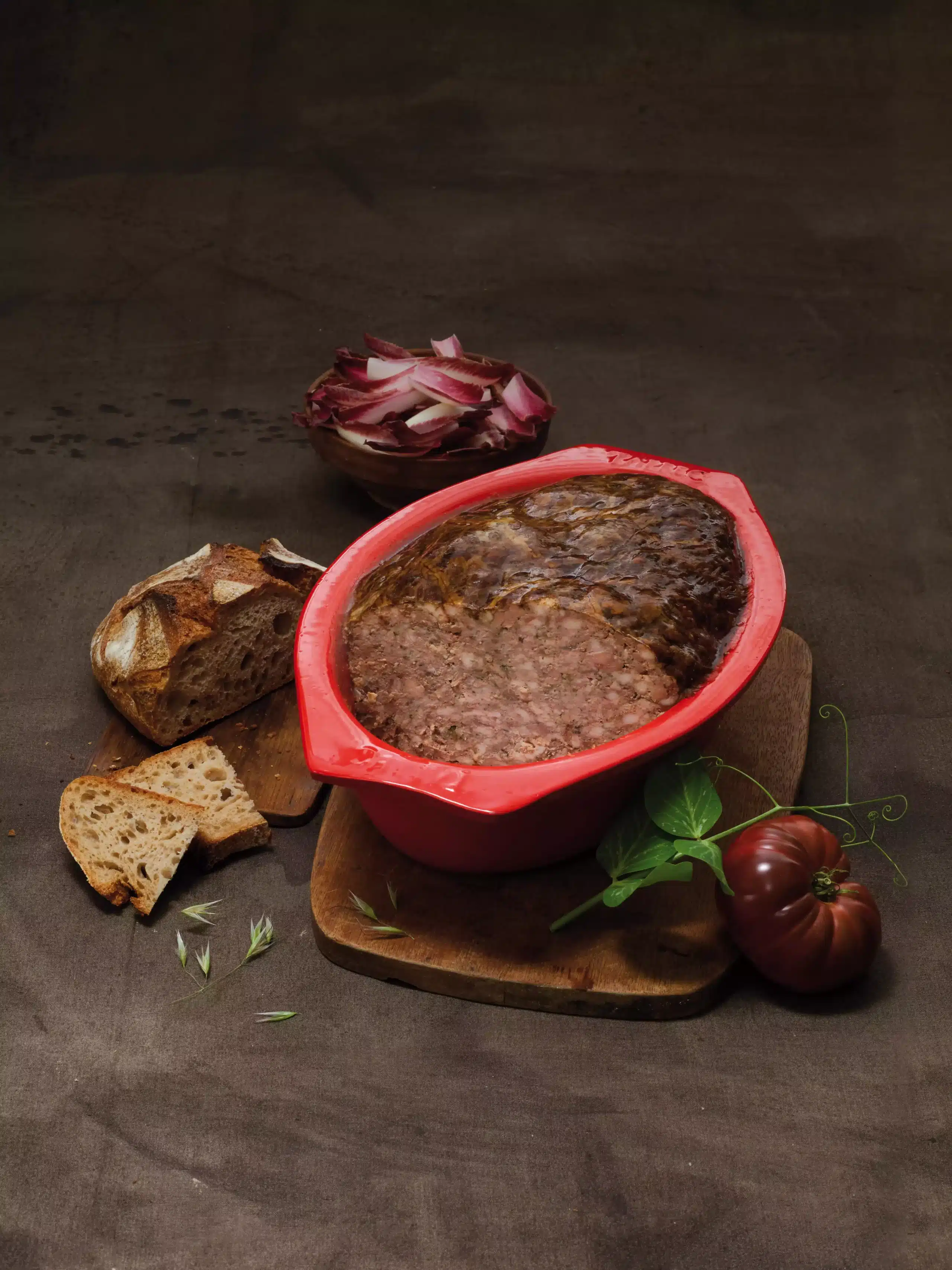 Terrine de campagne en céramique rouge sur planche en bois, entourée de pain artisanal, tomates et salade, sur fond brun