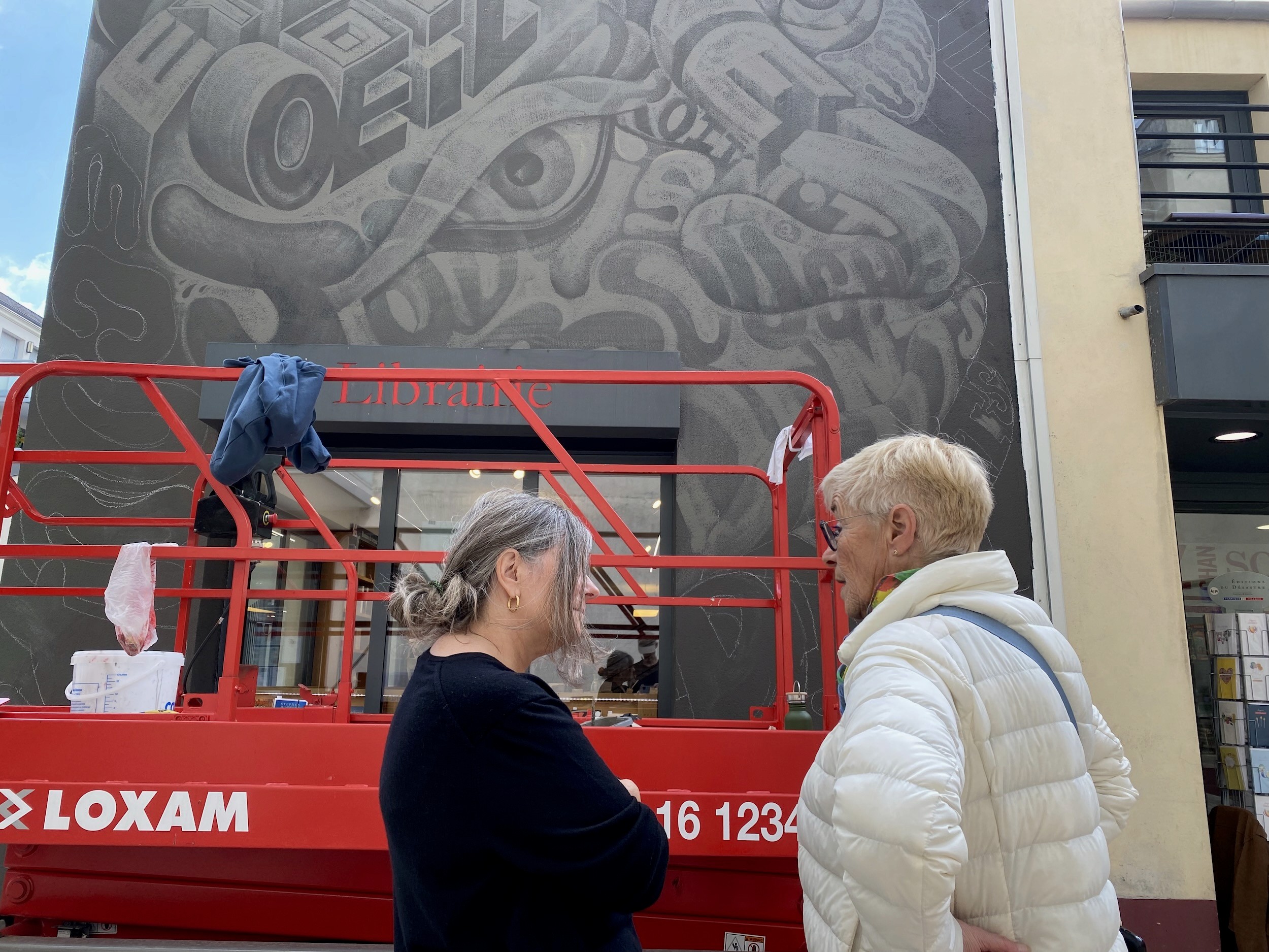 Two women talk in front of the bookshop wall, its grey lettering and an eye taking shape behind a red lift.