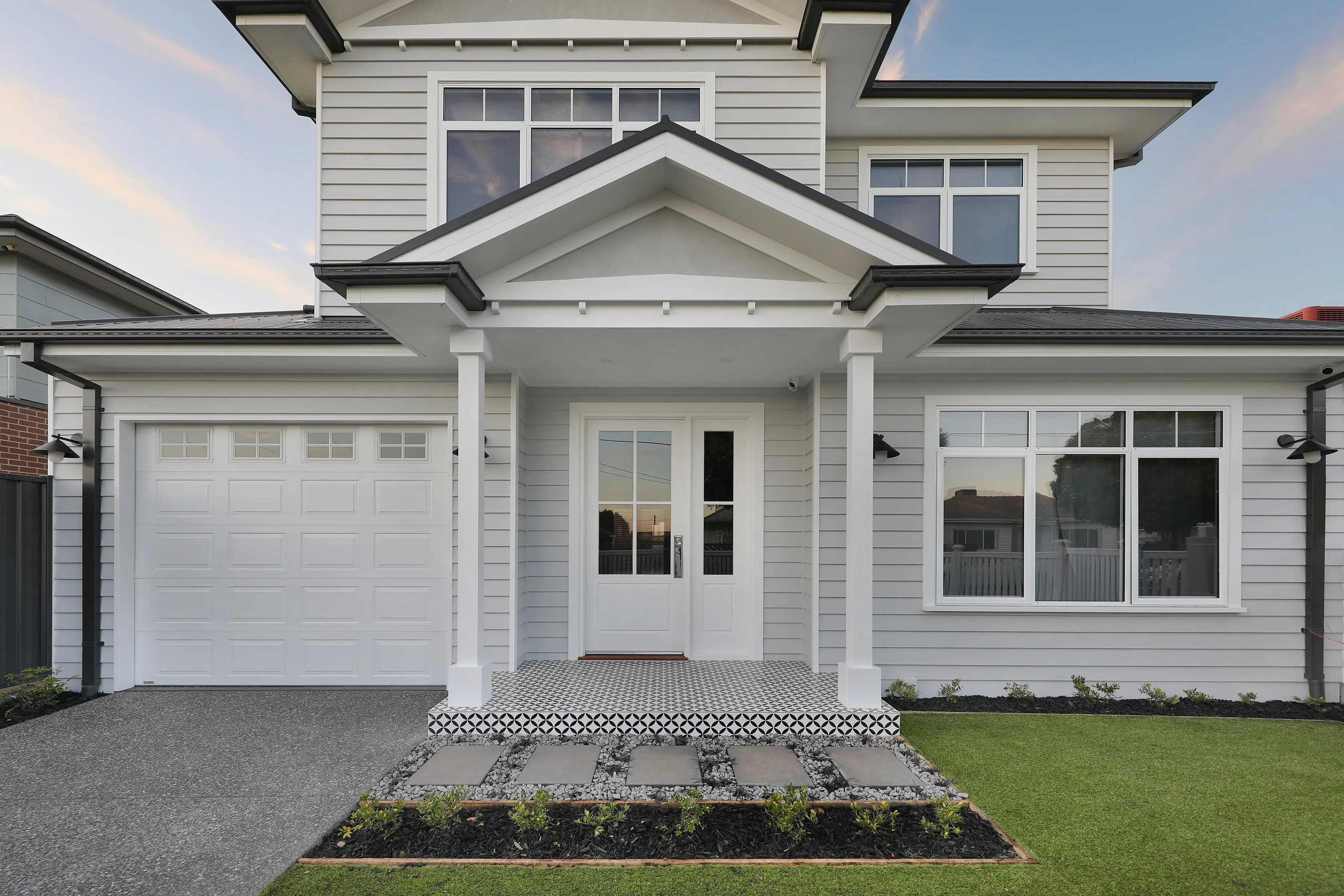 Entry portico of the Lalor Residence with a gabled roof, white columns, weatherboard cladding, a glazed white door with sidelight, patterned tile porch and a stepping-stone path
