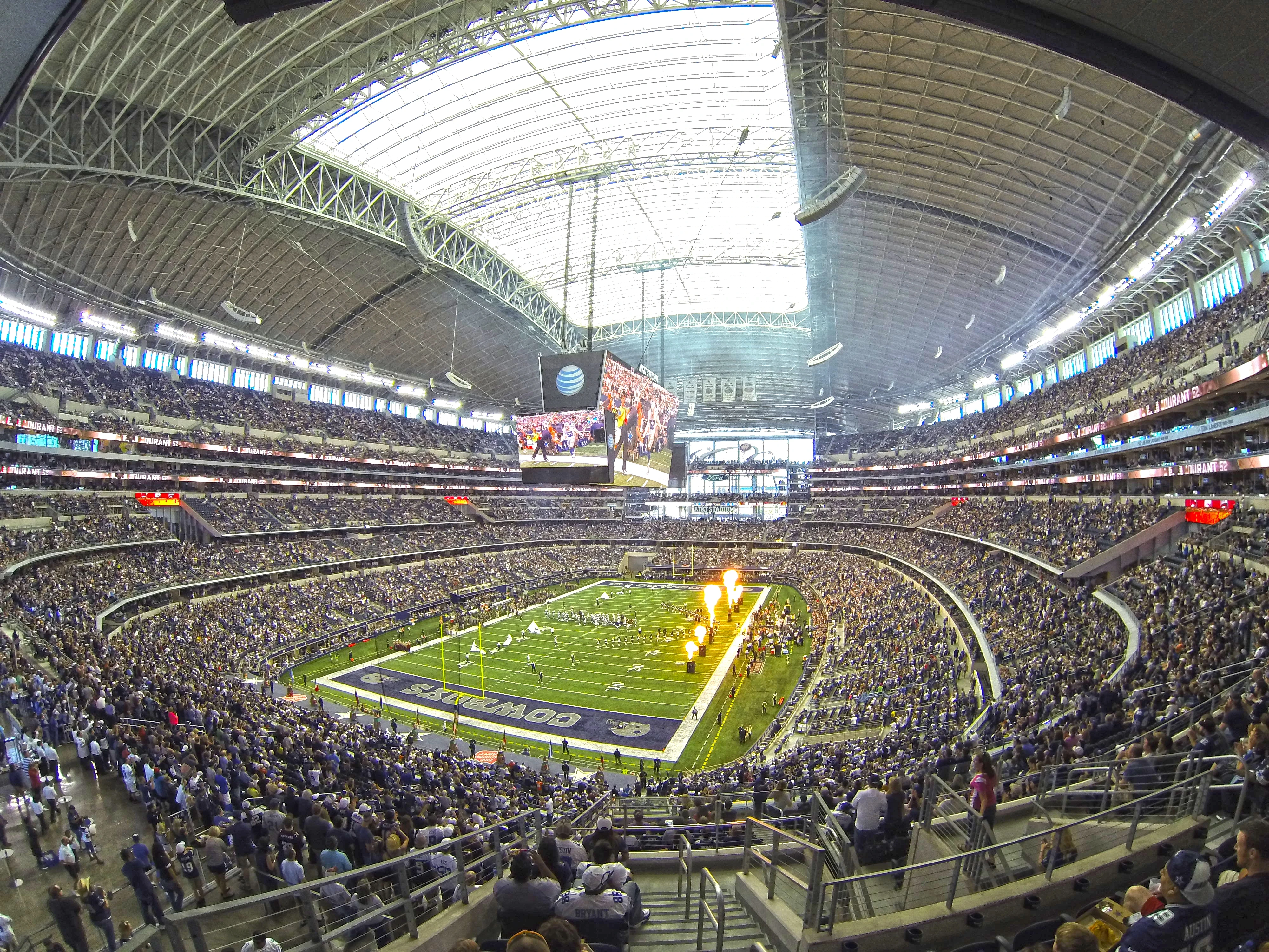 Packed NFL stadium interior during a Dallas Cowboys game with a massive center scoreboard and pyrotechnics on the field.