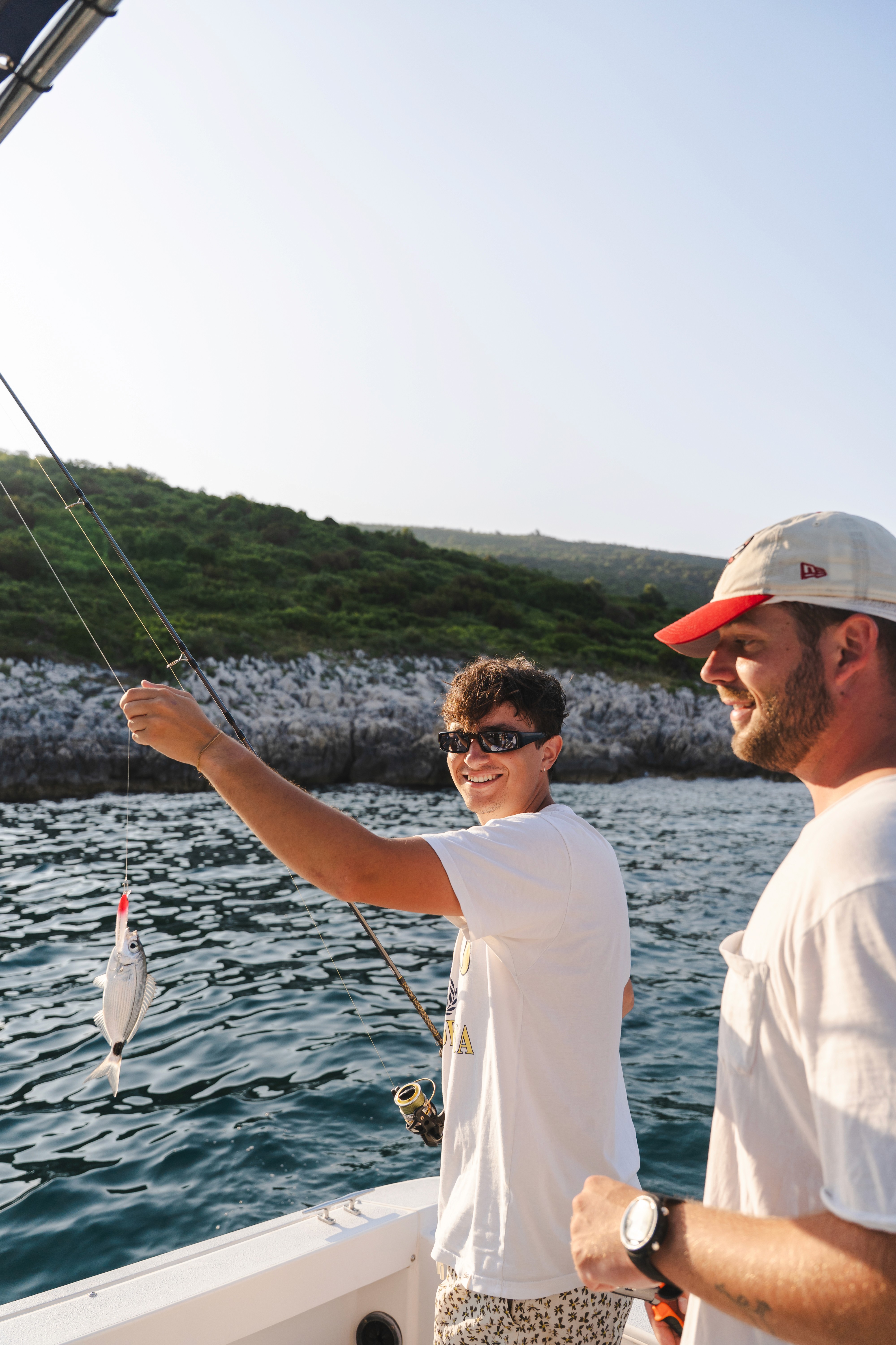 Two men enjoying a fishing trip on a boat, holding fishing rods and catching a fish on the Adriatic Sea.