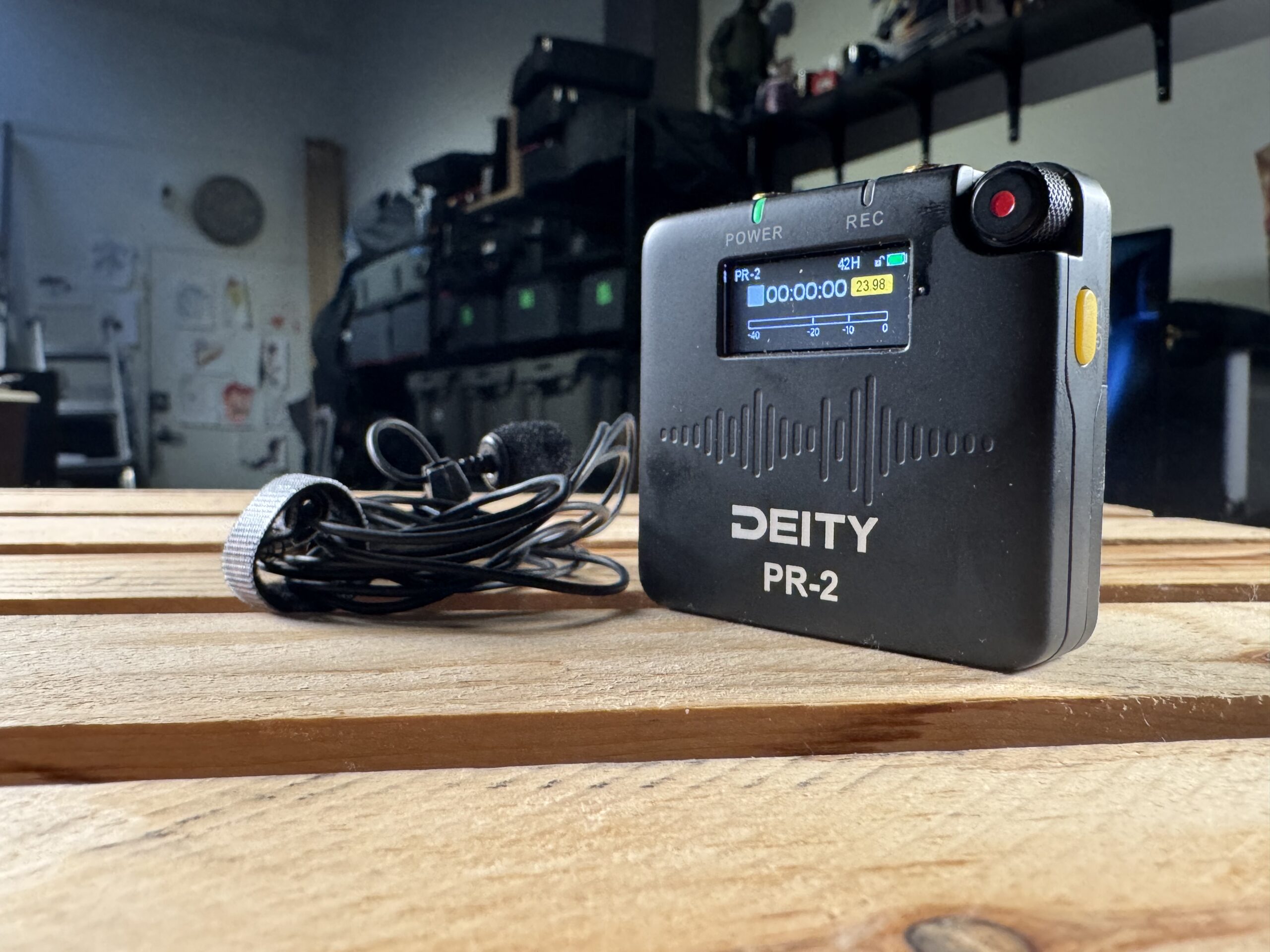 A Deity PR-2 pocket audio recorder with a coiled lavalier microphone sitting on a wooden surface in a studio.