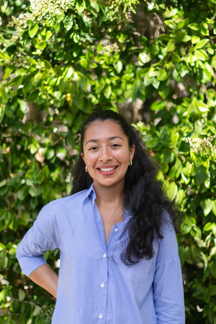 Uma mulher com cabelos longos e escuros sorri calorosamente enquanto está em frente a um fundo verde exuberante e cheio de folhas, vestindo uma camisa azul-clara de botões.