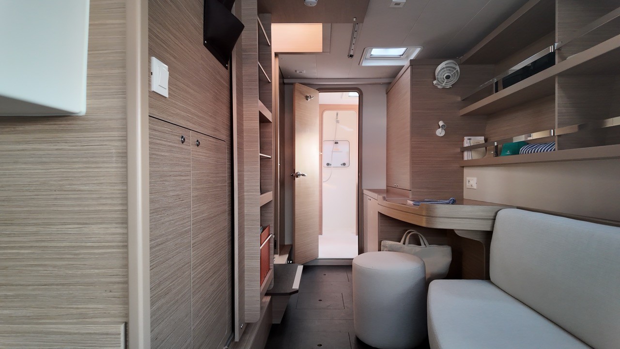 Interior view of a Lagoon 42 cabin corridor in warm wood tones, showing built-in shelving, a fold-out desk with stool, and an open door leading to the ensuite shower.