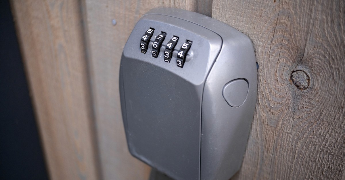 A gray combination key lock box with four numbered dials mounted on weathered vertical wooden boards.