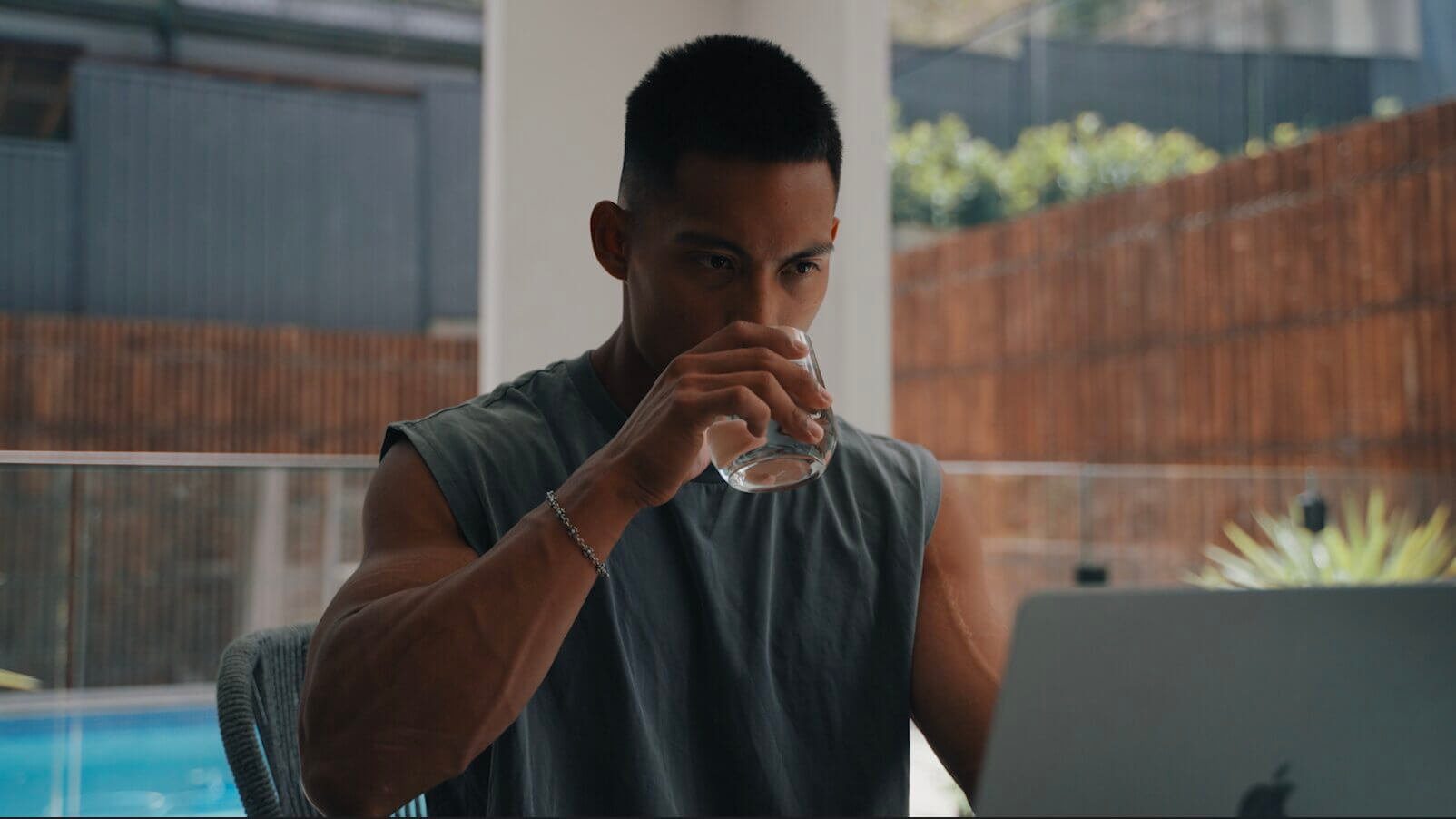 Man sips water, looking at a laptop by a pool.
