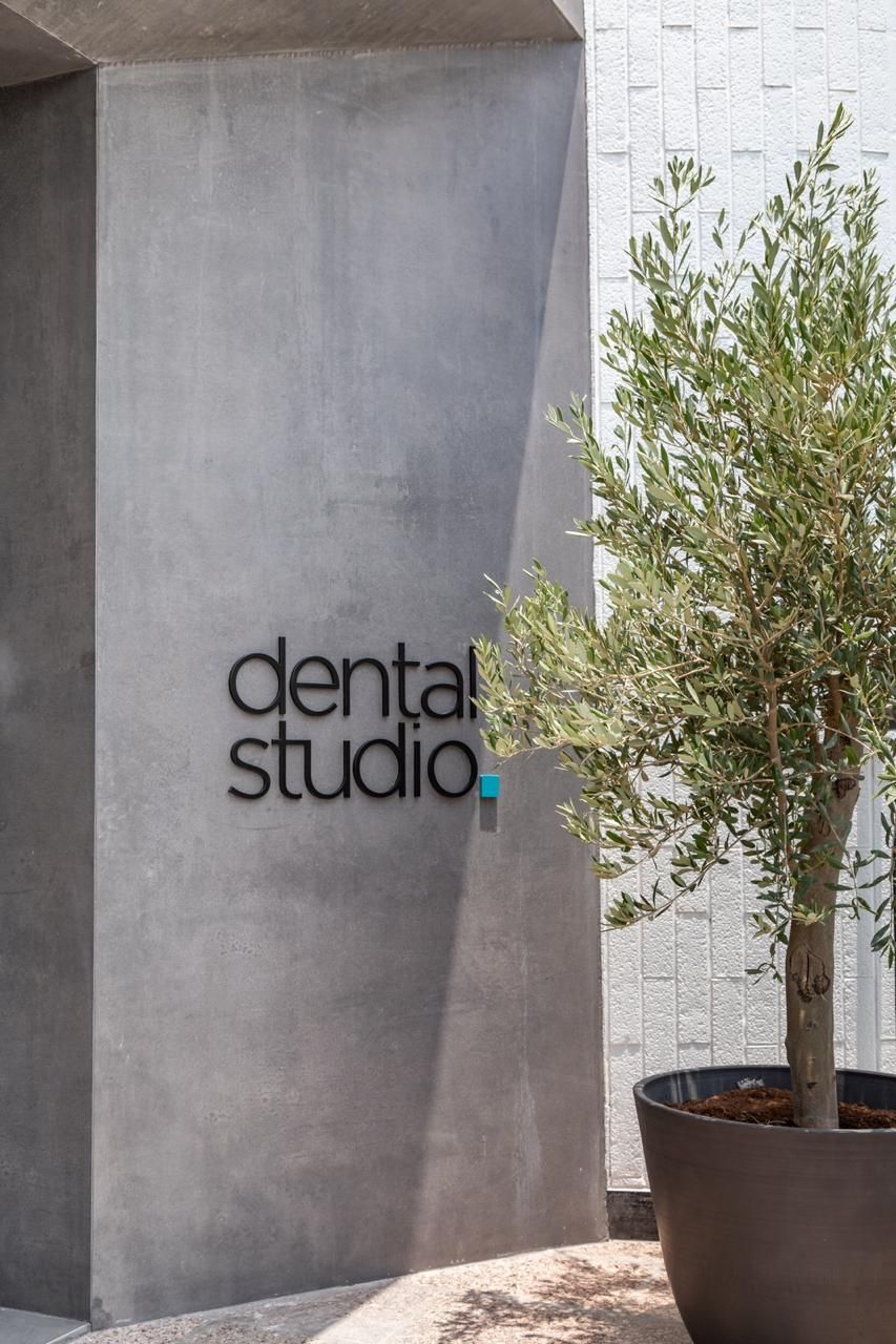 Exterior “dental studio” wall sign with minimalist black lettering mounted on a concrete clinic façade beside a potted tree.