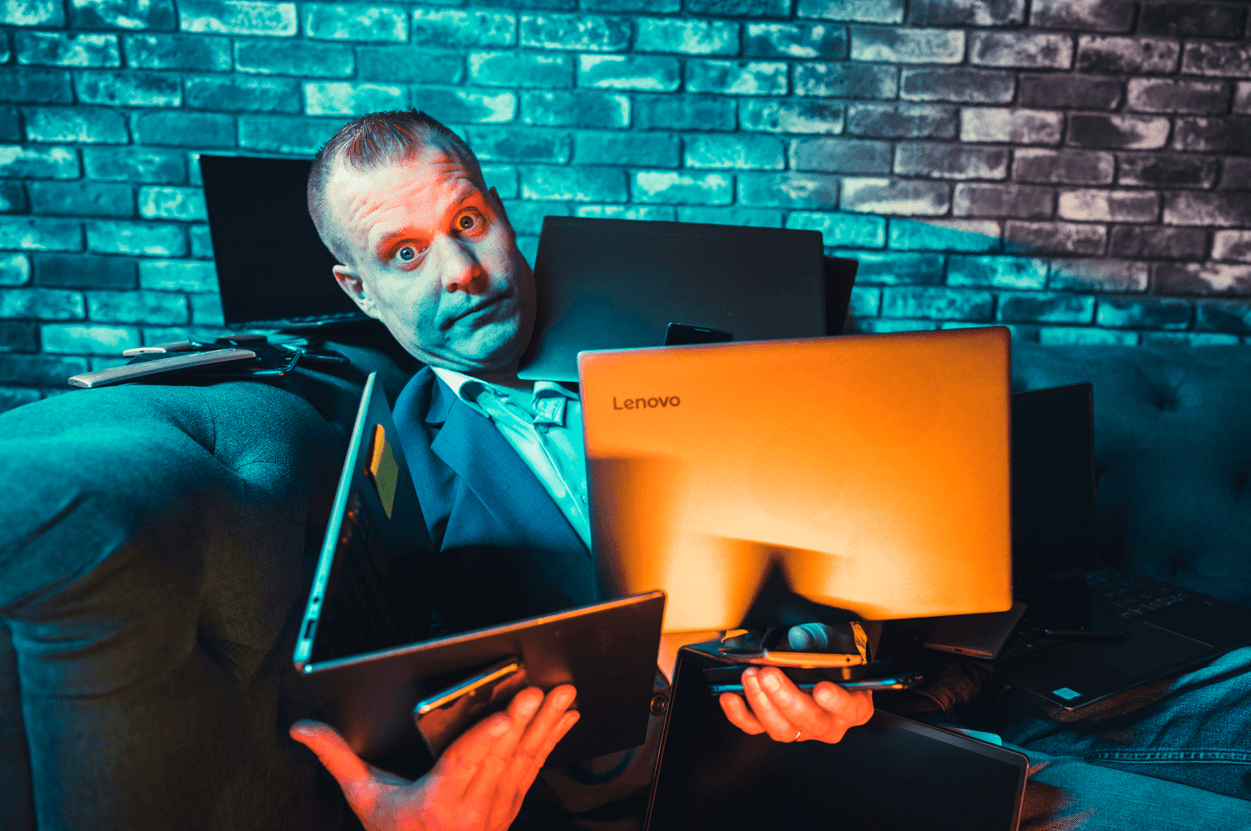 A man sits on a couch surrounded by laptops and other electronics, looking stressed and overwhelmed
