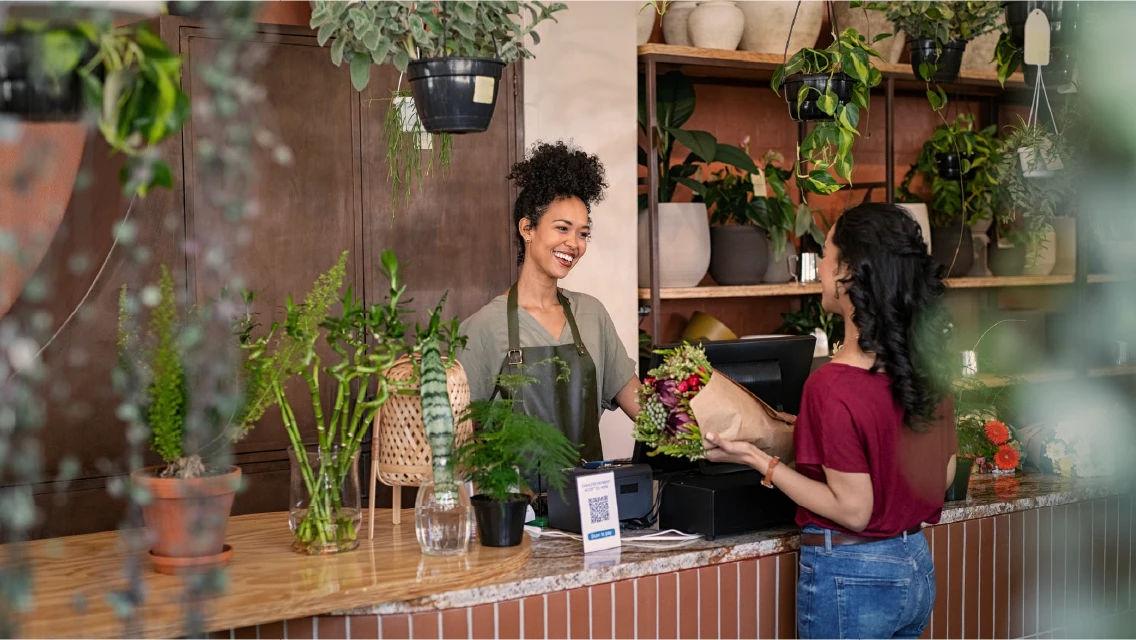 A smiling florist hands a wrapped bouquet to a customer at a plant-filled retail shop counter, illustrating an MFA alternative for retail workers without phones.