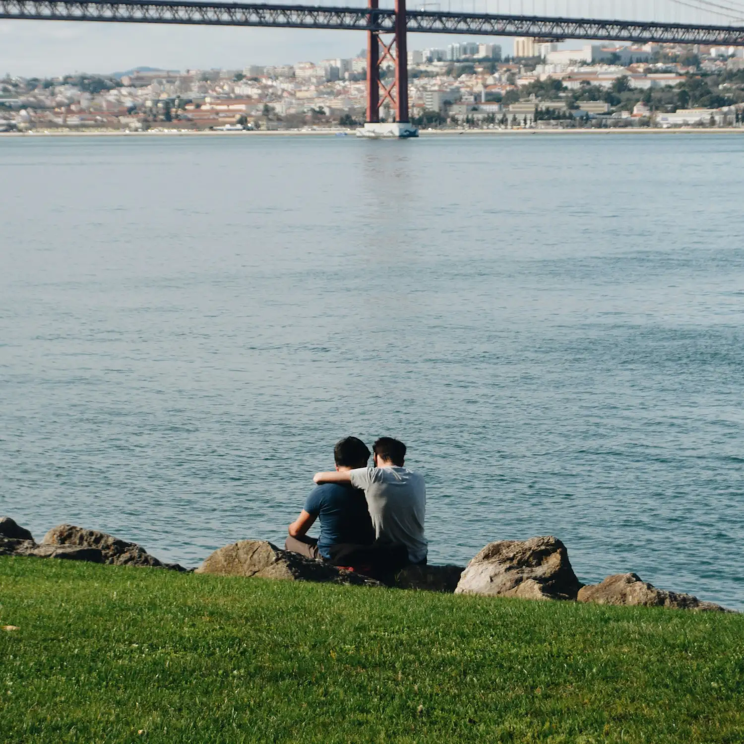 Two people sitting together by the water, embracing while overlooking a city bridge, symbolizing connection, support, and gay-affirming therapy in NYC.