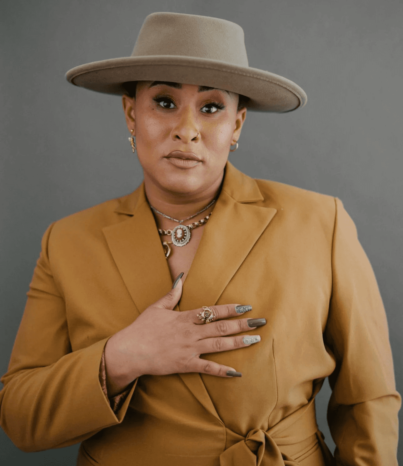 A person in a tan outfit and wide-brimmed hat poses with a hand over their heart against a neutral background.