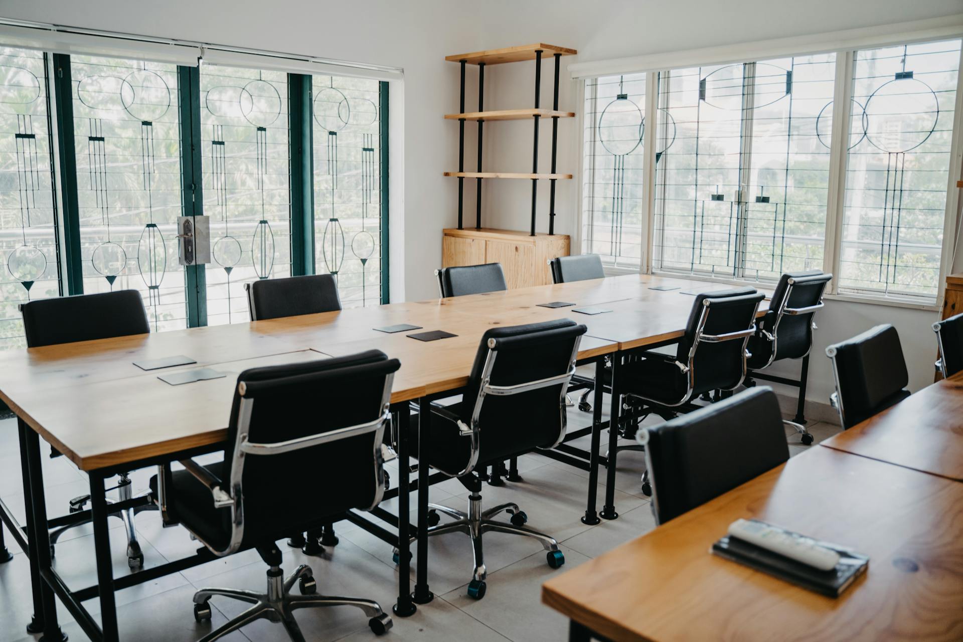 A bright, modern meeting or conference room with large windows providing ample natural light. The room features a long, light wooden table surrounded by several black executive-style rolling chairs. A simple wooden shelving unit with black metal supports is visible against the far wall.