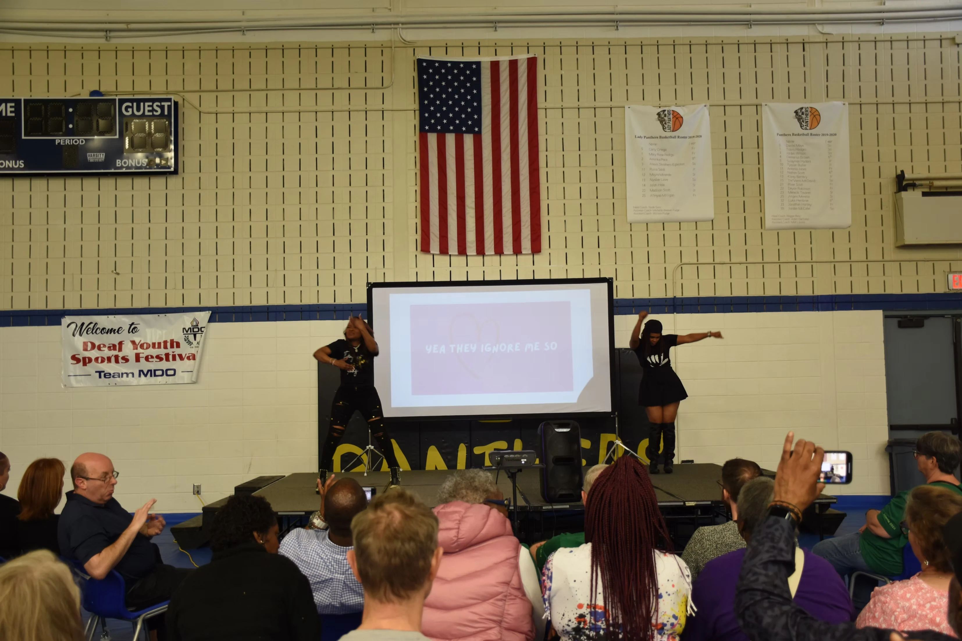Two women in black perform on a stage in a gymnasium while an audience watches a presentation.