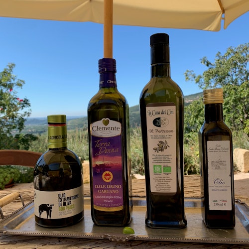 Cuatro botellas de aceite de oliva sobre una bandeja al aire libre, con un paisaje escénico de árboles y un cielo despejado bajo una sombrilla de patio.