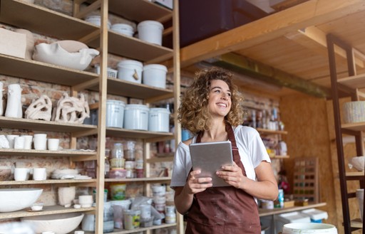 A smiling person stands in a shop surrounded by shelves stocked with various ceramic dishes and pottery.