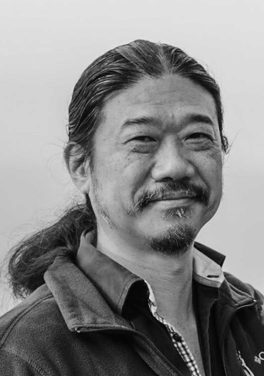 Smiling person with curly hair, wearing a black shirt, against a warm, dark background.