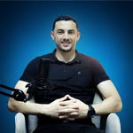 Young person with wavy hair poses confidently in sunlight against a modern building backdrop. Wears a black blazer and silver necklace, conveying elegance.