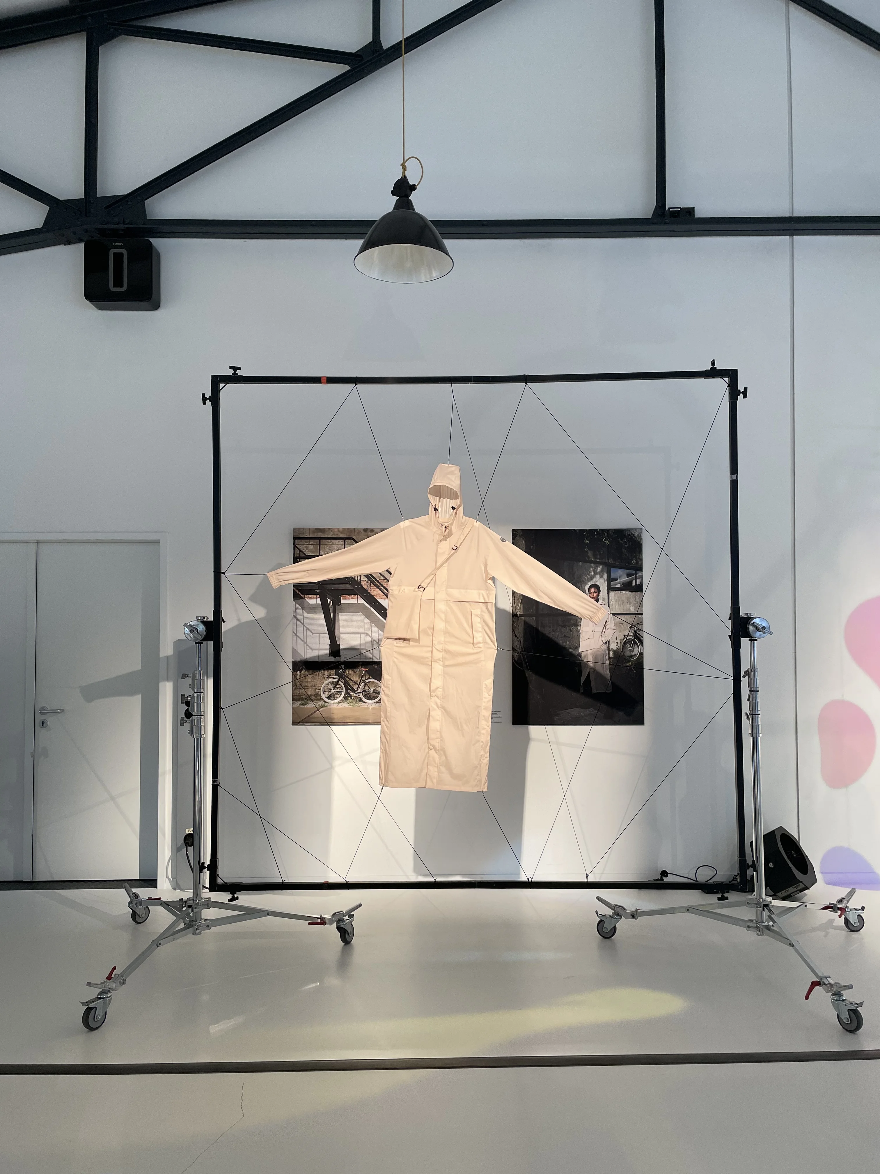 Beige AGU rain coat suspended in a tensioned wire frame in front of photo prints under an industrial pendant lamp.