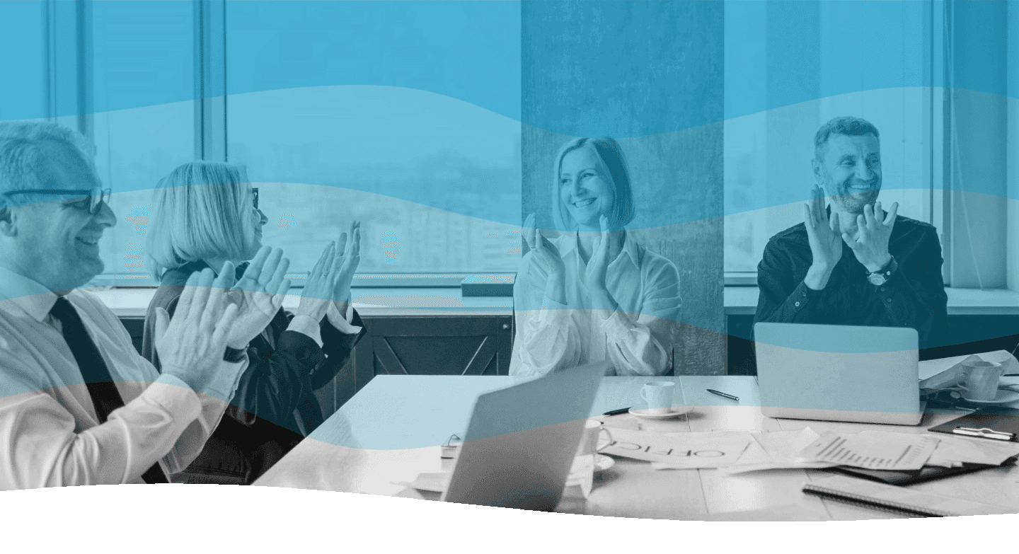 A group of professionals in an office setting sit around a table with open laptops and scattered paperwork, smiling and applauding each other, suggesting a collaborative and successful meeting.