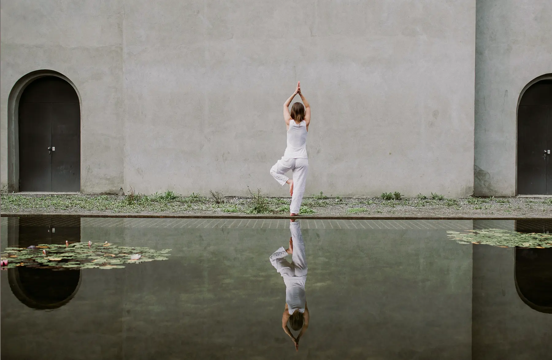 A person practices yoga in a serene outdoor setting, standing near a tranquil reflecting pool with a minimalist stone wall in the background, embodying wellness and recovery at a premium spa-like environment.