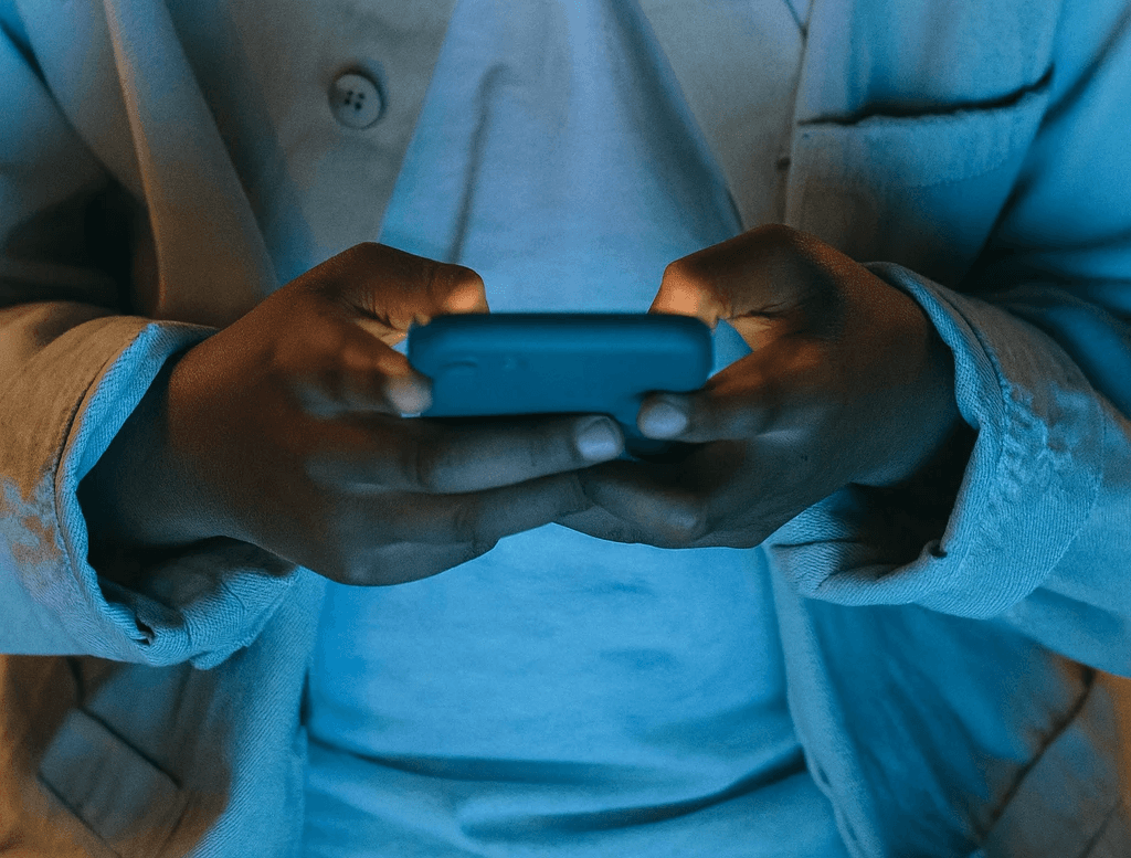 Garoto segurando celular, a imagem está um pouco escura e o celular ilumina a roupa da pessoa