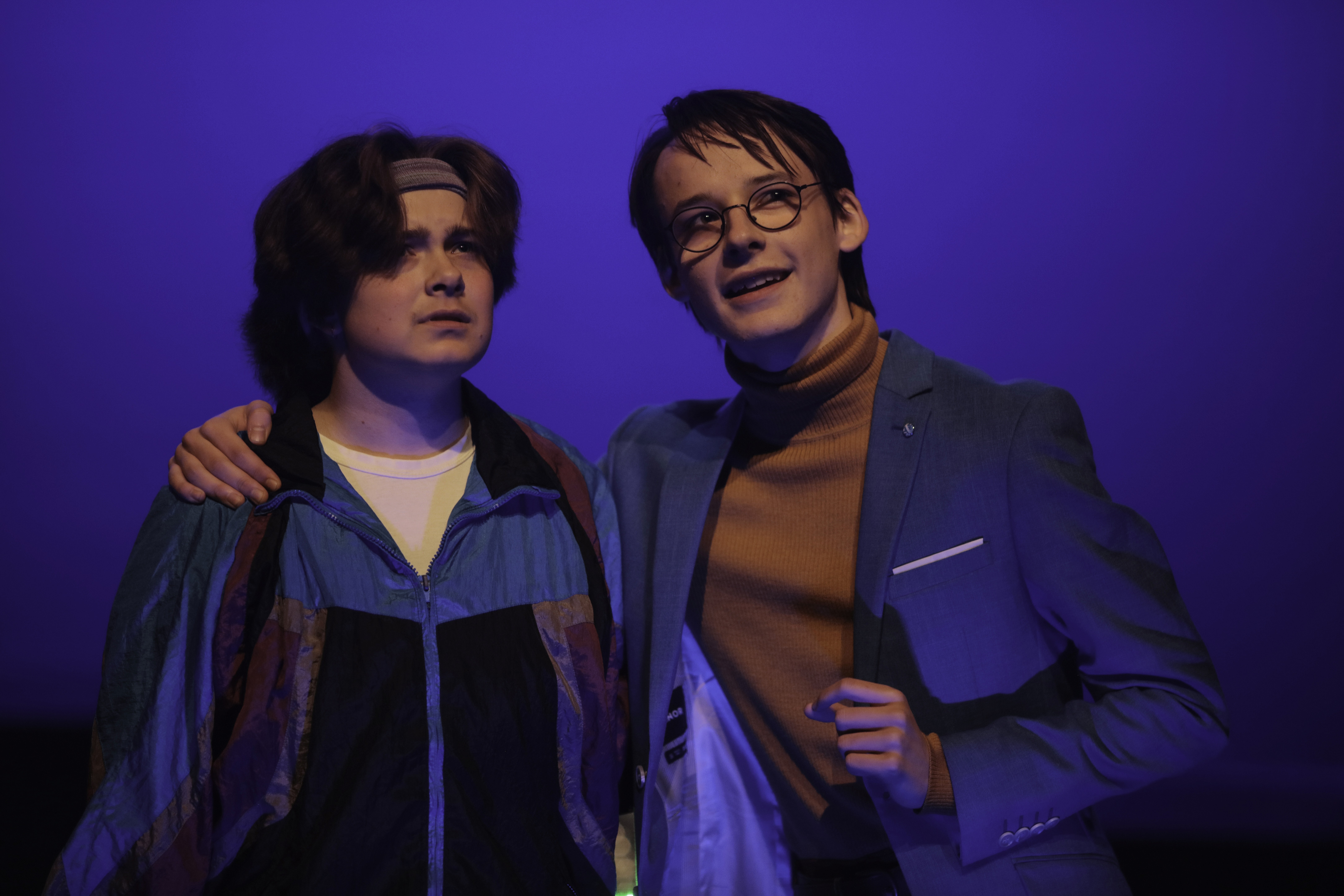 A teen in a retro windbreaker stands beside a confident character in a teal blazer and glasses, who has an arm around their shoulder. Both look ahead under cool blue lighting, as if witnessing something extraordinary.