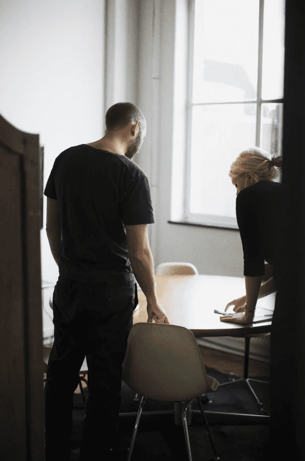 Woman in a tailored beige suit holding a structured brown leather handbag, standing in front of a softly lit modern background.