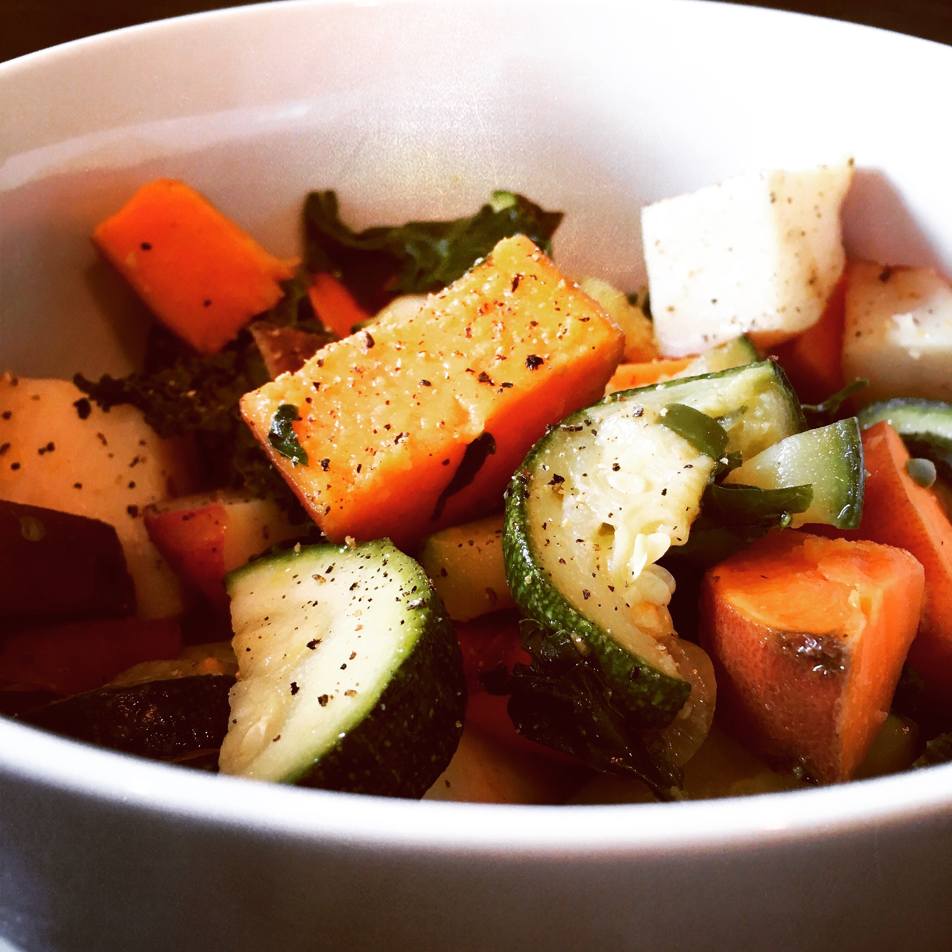 A white bowl filled with a vibrant medley of roasted vegetables, including zucchini, carrots, and sweet potatoes, seasoned with black pepper.