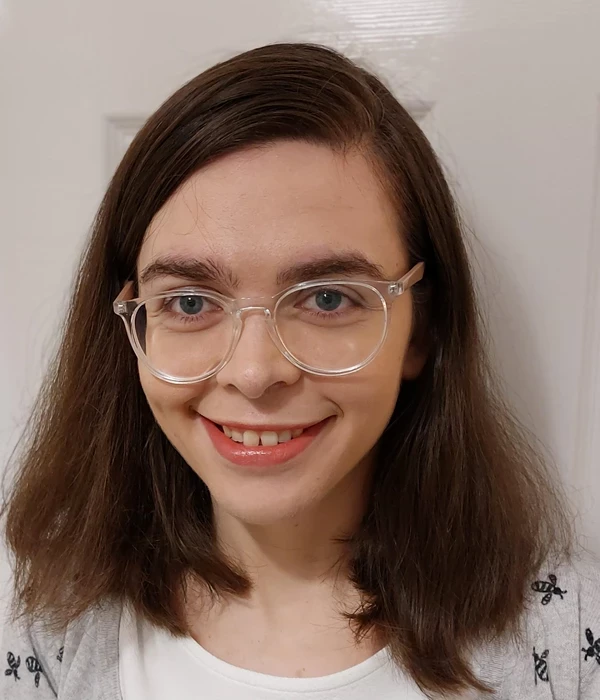 A person with shoulder‑length brown hair wears glasses and a light‑coloured top with a subtle pattern. The photo is taken against a plain indoor wall, giving it a straightforward, portrait‑style composition.