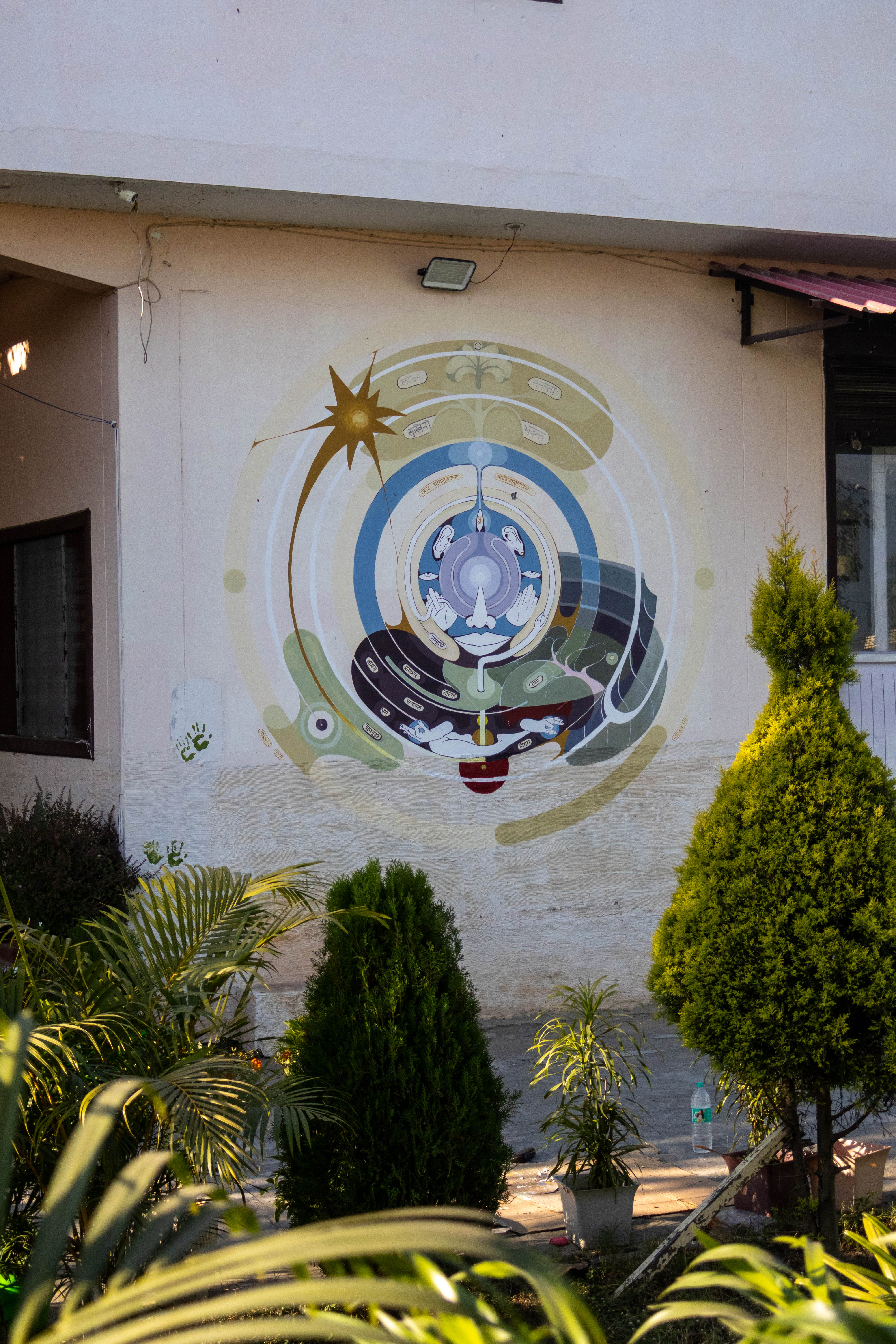 A mandala-style mural with spiritual symbols painted on a white wall, surrounded by green shrubs.