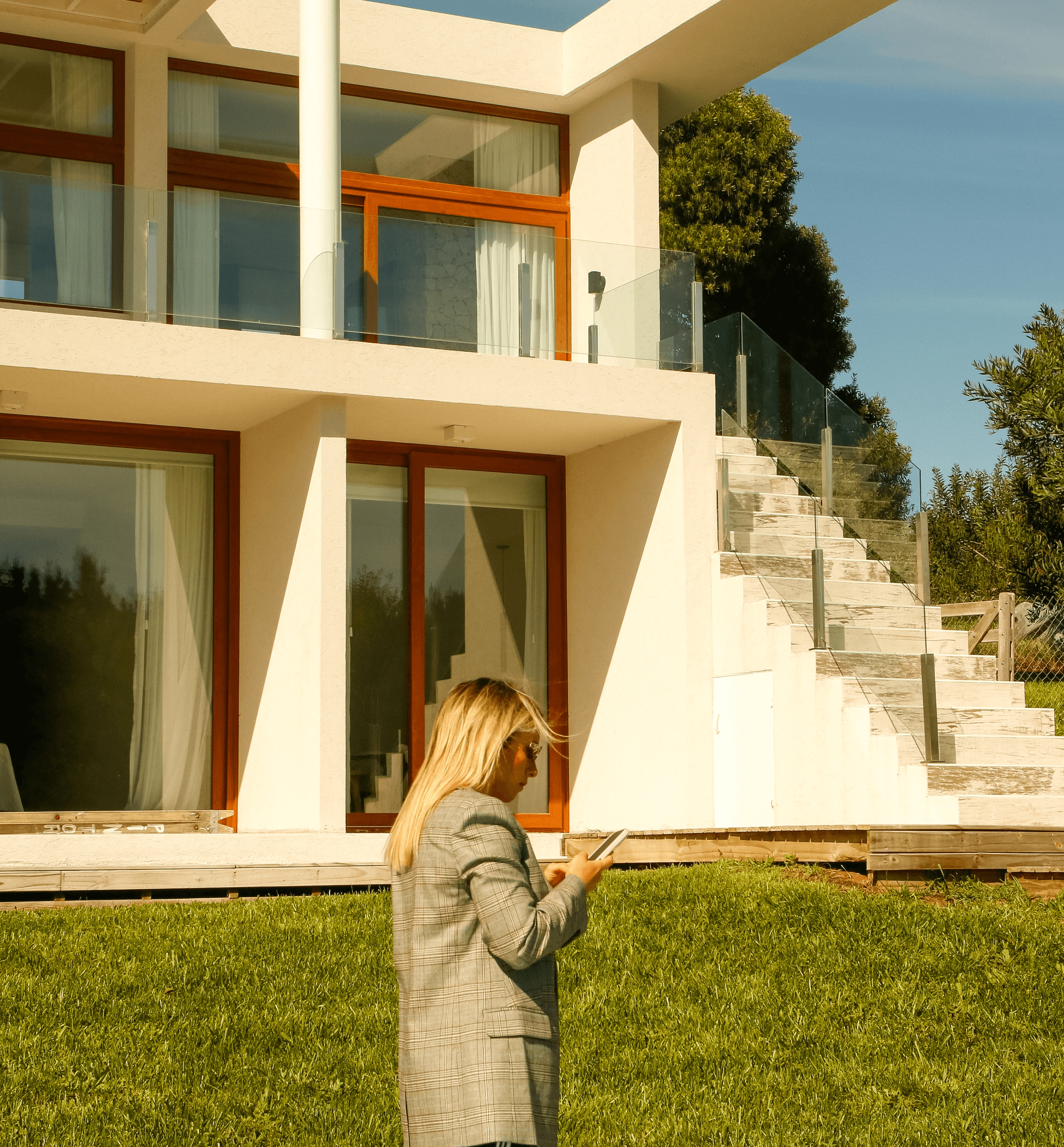 Mujer con blazer a cuadros consulta su celular en jardín, frente a casa moderna de dos plantas con escaleras y barandas de vidrio.