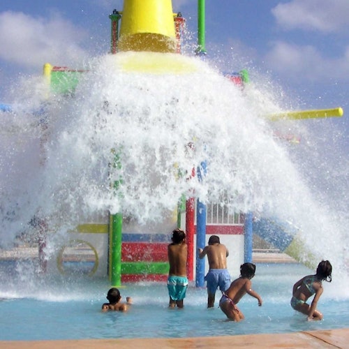 Niños jugando bajo un gran chorro de agua de una colorida estructura de juegos acuáticos en una piscina en un día soleado.