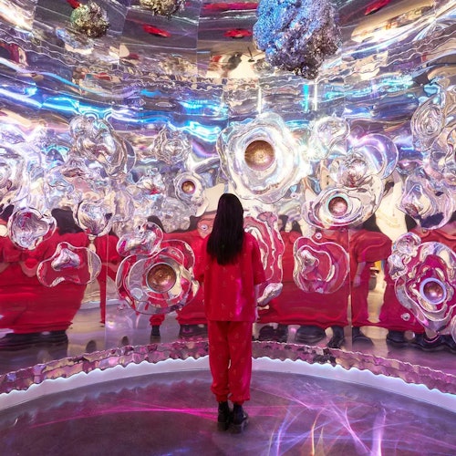Una persona vestida de rojo está de pie dentro de una instalación de arte inmersiva y reflectante, con formas abstractas y metálicas en las paredes y el techo.