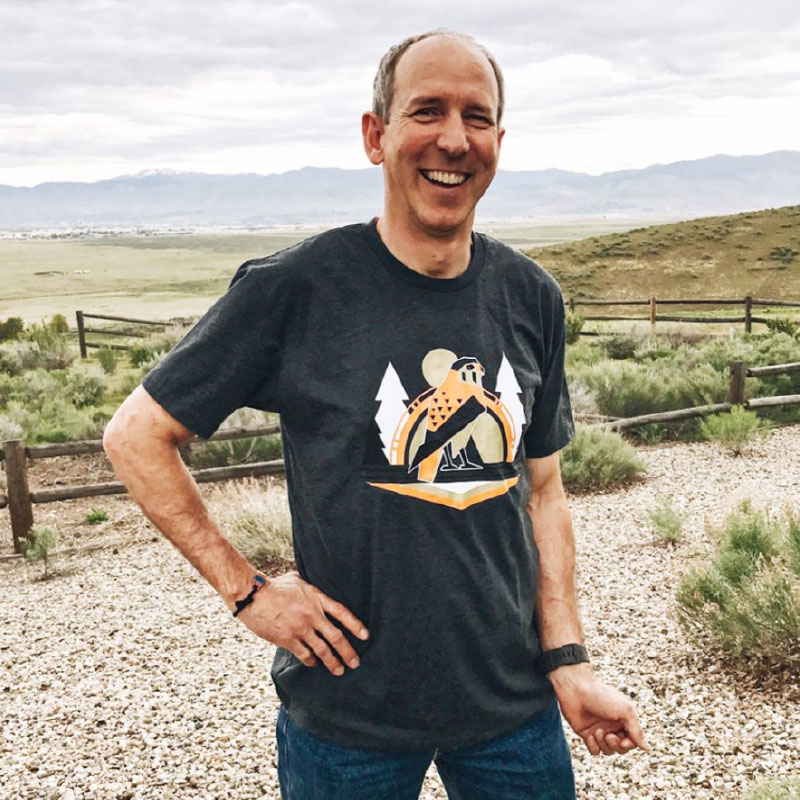Smiling man wearing a dark gray t-shirt with a nature-inspired design, standing outdoors in a scenic landscape.