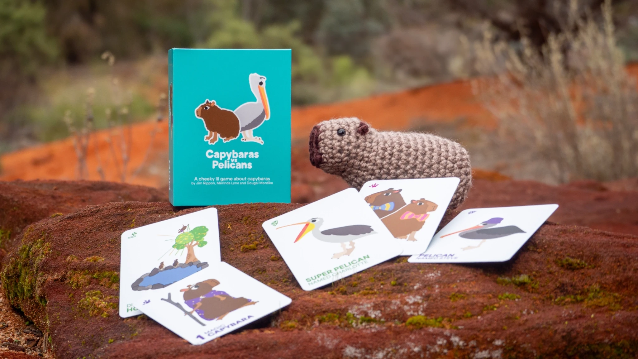 Capybaras vs Pelicans board game box resting on a rock alongside a crochet capybara plushie and five playing cards.