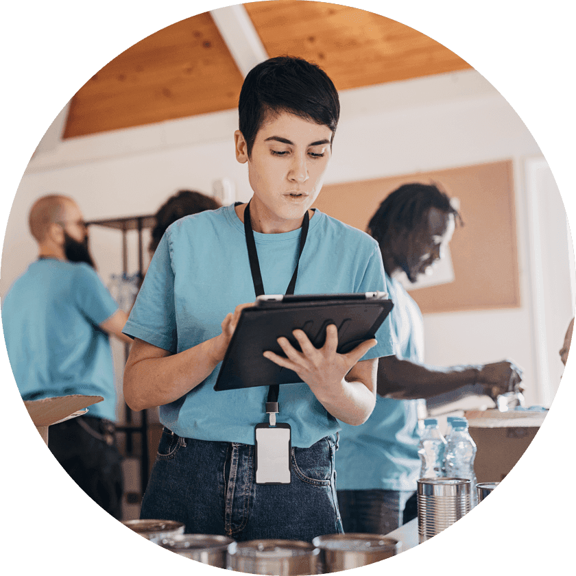 A young woman in a blue shirt holding a clipboard while concentrating on her task. In the background, other volunteers are organizing items on tables, working in a bright room.