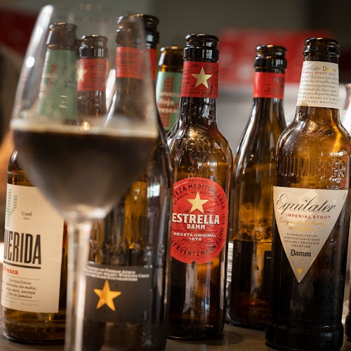 前景中有一个装满饮料的玻璃杯，旁边放着几瓶带有不同标签的啤酒，包括Estrella Damm和Equilater。