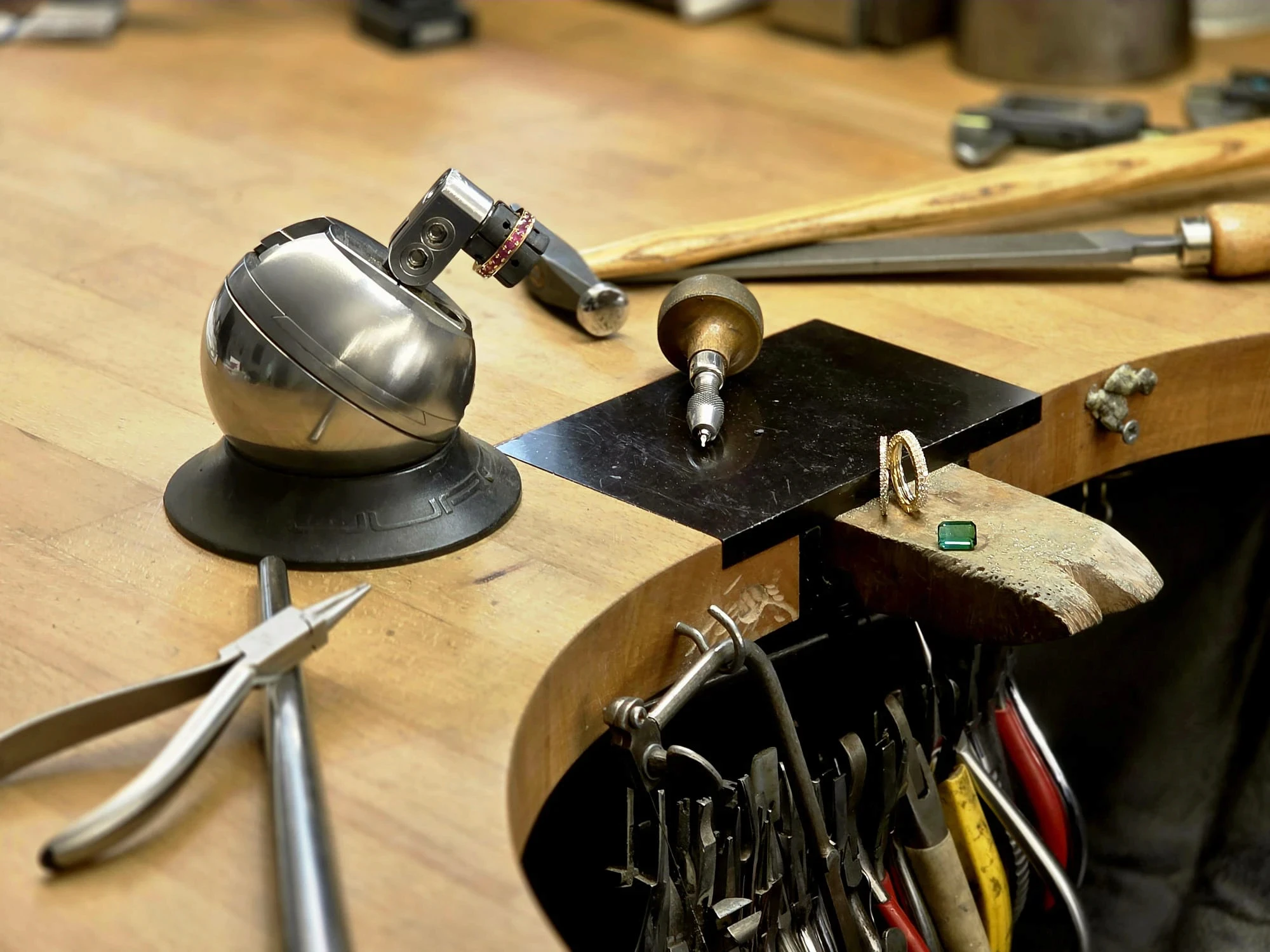 Nahaufnahme einer detaillierten Werkbank in einer Goldschmiedewerkstatt mit Werkzeugen wie Zangen, Feilen und einer Gravurkugel. Auf einer Arbeitsplatte aus Holz liegen ein Goldring, eine Gravurnadel und ein grüner Smaragd.