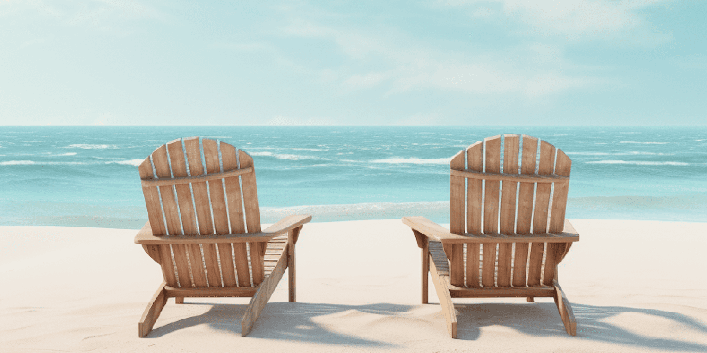 Two wooden lounge chairs facing the calm blue ocean on a sunny sandy beach.