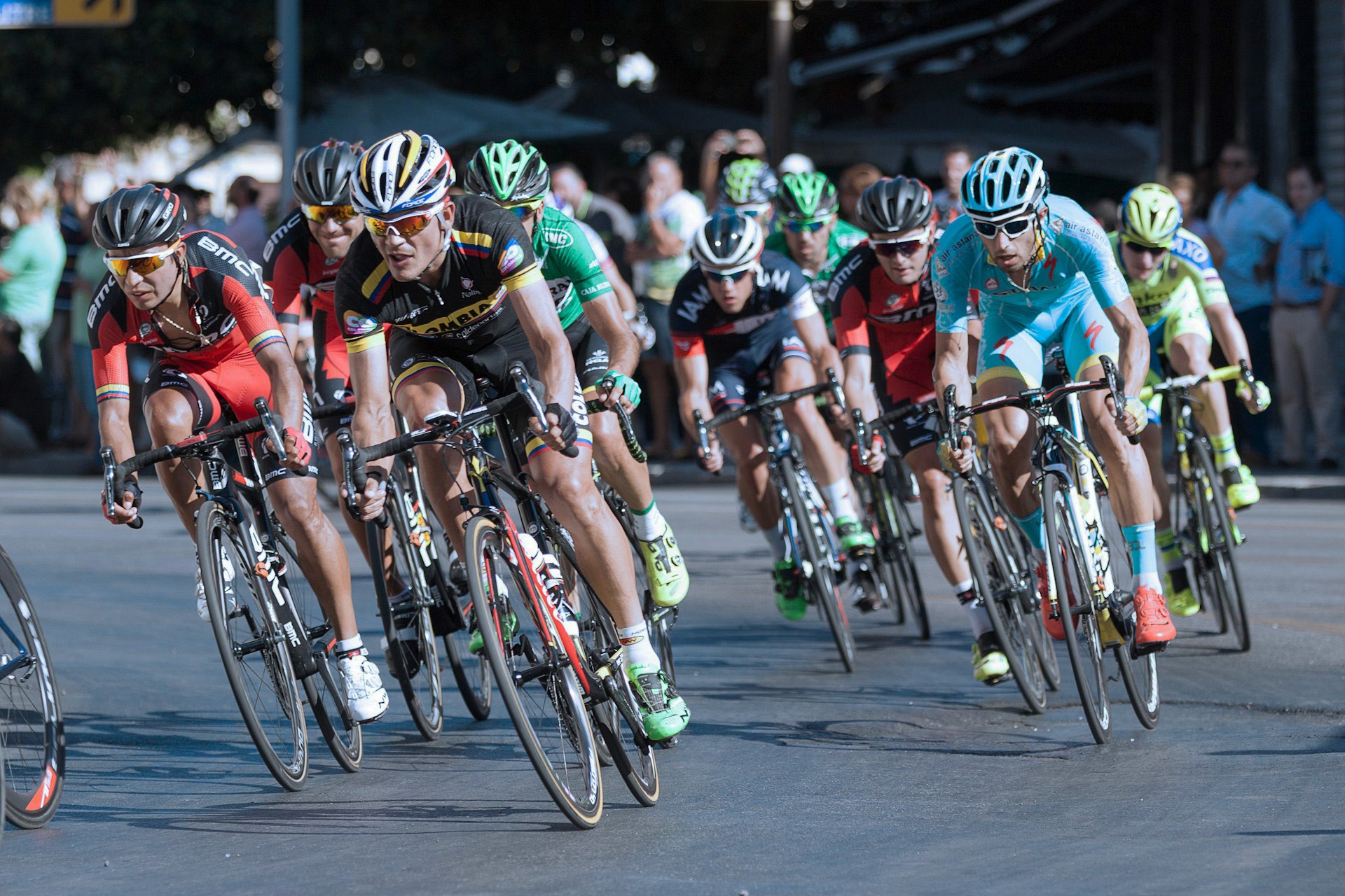 A peloton of professional cyclists in helmets and team jerseys race tightly together on a city street.