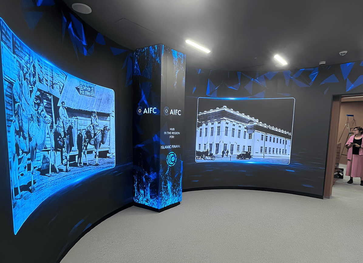Curved, dark exhibition room with blue geometric light effects and two large wall screens showing black-and-white archival photos; a central glowing LED pillar displays the AIFC logo, and a person stands in an open doorway on the right.
