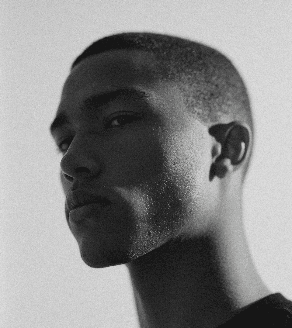 Black and white editorial portrait of a man in profile with strong side lighting, soft grain, and minimalist studio background