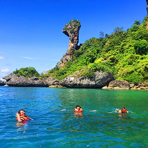 Three people swim in clear blue water near a tall rock formation and lush green cliffs under a bright blue sky.